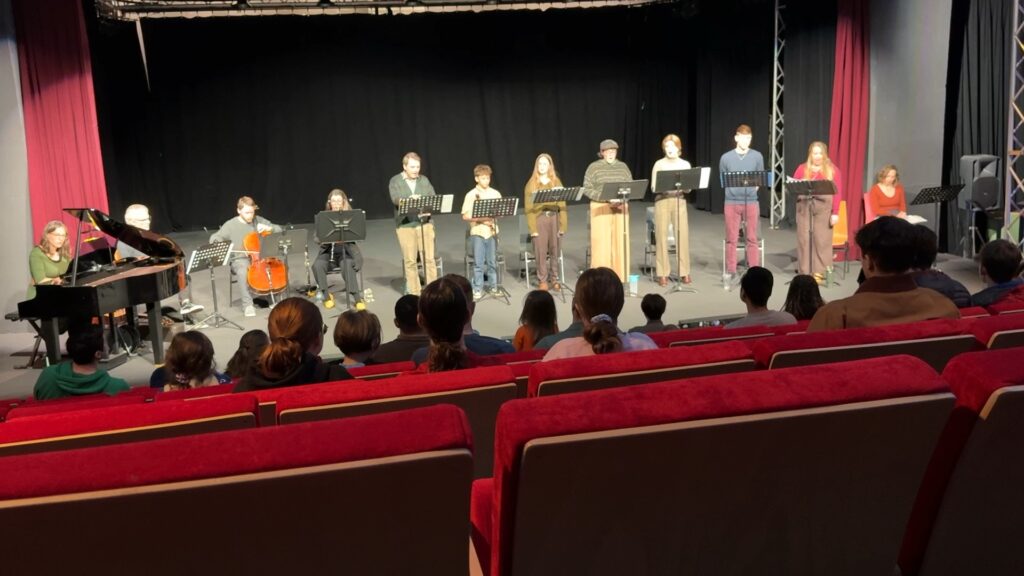 The workshop cast of Piper: A New Musical performs on stage with music stands. Composer Avril Callicutt sits at the piano